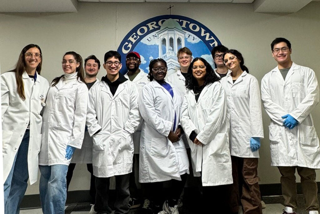 Biotech students wearing lab coats and gloves stand in a hallway in front of a mural of the Medical and Dental Building cupola